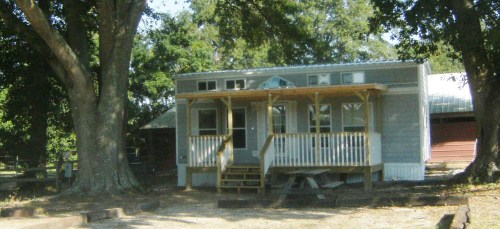 A small, light blue house with a front porch, surrounded by trees and a gravel yard.