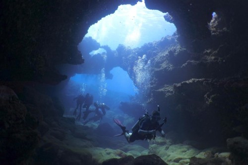 Divers explore an underwater cave, surrounded by rocky formations and bubbles in a serene blue environment.