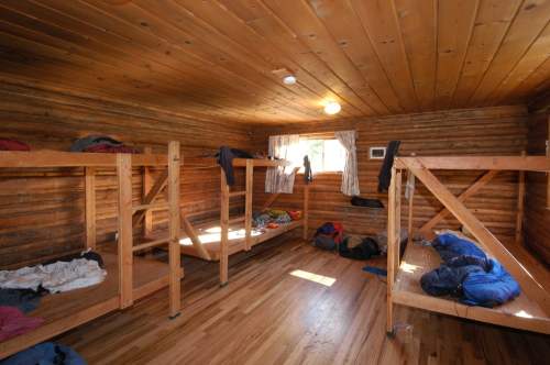 Interior of a rustic wooden cabin with bunk beds, scattered clothing, and a window letting in natural light.