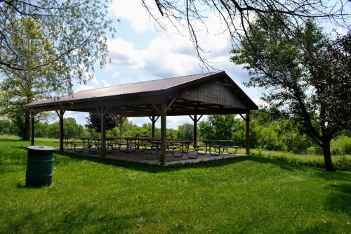 A shaded picnic shelter with tables, surrounded by green grass and trees under a partly cloudy sky.