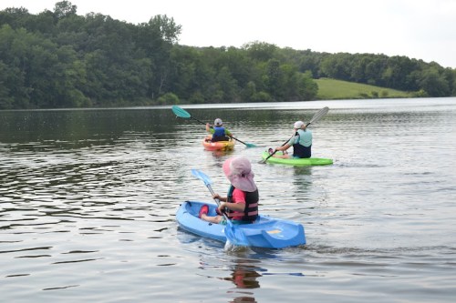 Three people kayaking on a calm lake surrounded by greenery under a cloudy sky.
