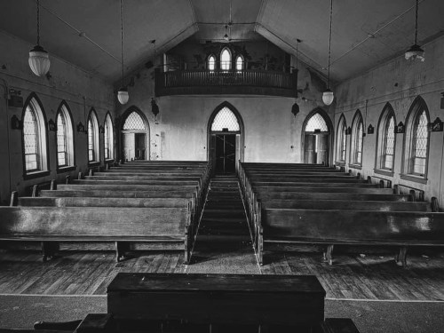 Interior of an empty church with wooden pews, arched windows, and a simple altar, captured in black and white.
