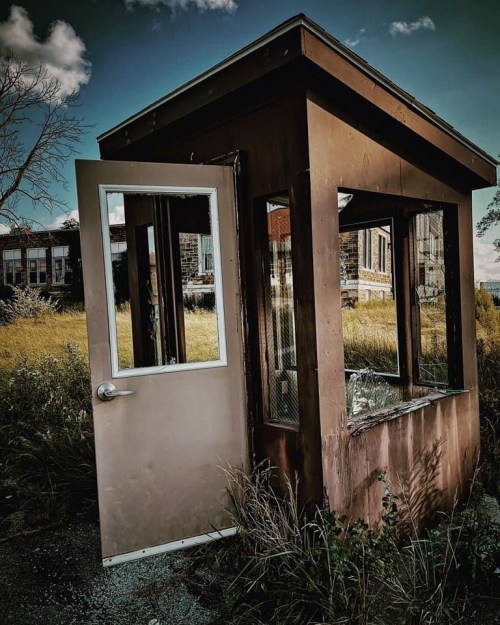 An abandoned, weathered booth with a glass window, surrounded by overgrown grass and a distant building.