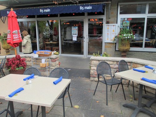 Outdoor dining area of a restaurant with empty tables, blue napkins, and a welcoming entrance.