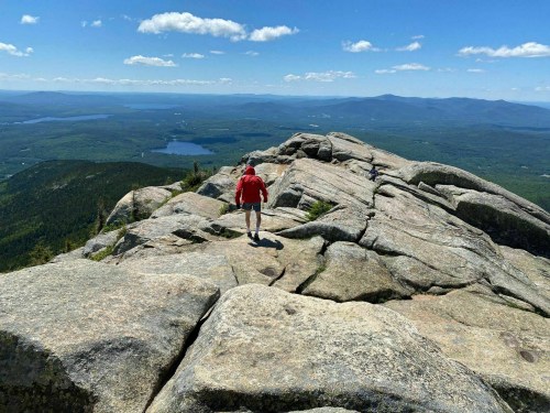A person in a red jacket walks on rocky terrain, overlooking a vast landscape with lakes and mountains in the distance.