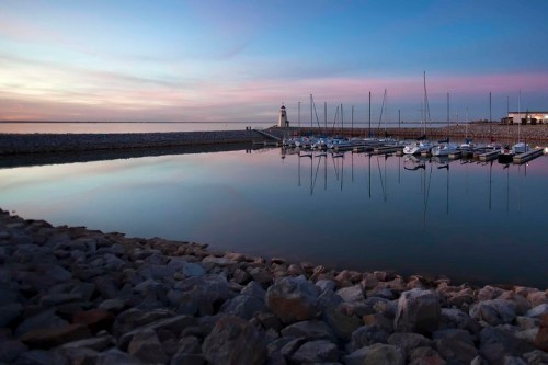 A tranquil marina at sunset, with boats reflected in calm water and a lighthouse in the distance.