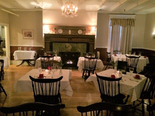 Cozy restaurant interior with white tablecloths, black chairs, and a fireplace, softly lit by a chandelier.