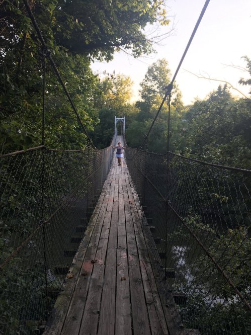 A person walks across a wooden suspension bridge surrounded by lush greenery and trees.