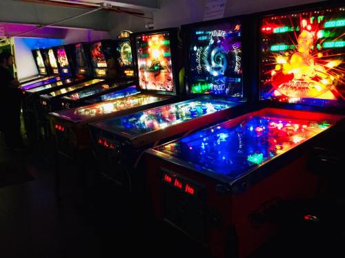 A row of colorful pinball machines with bright lights in a dimly lit arcade setting.