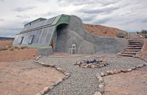 A modern, eco-friendly home with solar panels, surrounded by rocky terrain and a fire pit, under a cloudy sky.