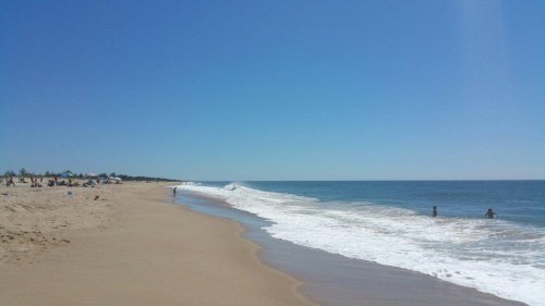 A sunny beach scene with gentle waves, a clear blue sky, and a few people walking along the shore.