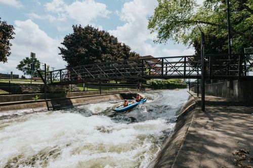 A person kayaking on a river with a bridge overhead and trees lining the banks under a partly cloudy sky.
