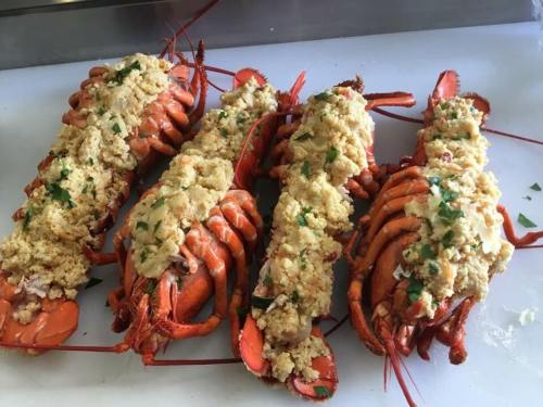 Four cooked lobsters topped with a golden breadcrumb stuffing and garnished with parsley, arranged on a white surface.