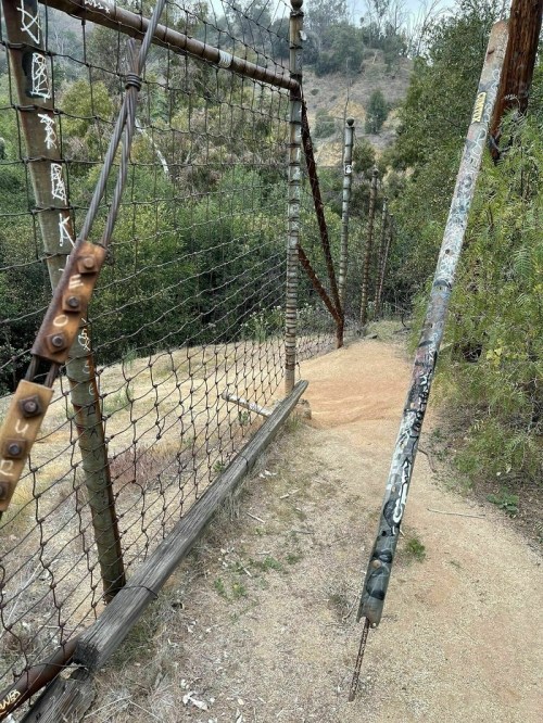 A weathered fence with a broken post, surrounded by greenery and a dirt path leading into the distance.