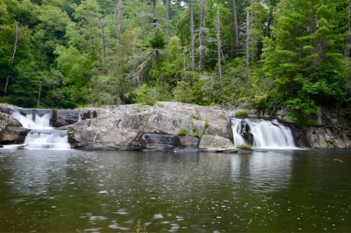 A serene landscape featuring a calm river with cascading waterfalls, surrounded by lush green trees and rocky formations.