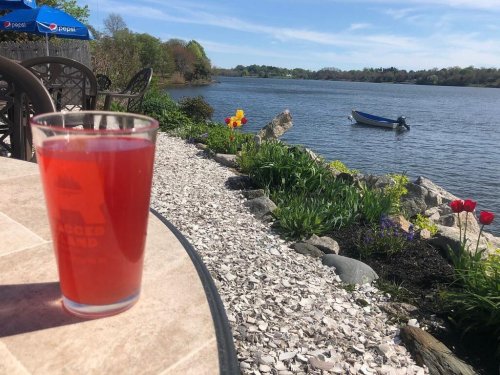 A glass of pink drink on a table by a scenic river, with flowers and a small boat in the background.