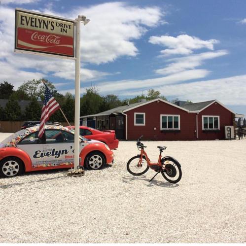 A colorful drive-in restaurant with a sign, a parked car, and a bicycle in a gravel lot under a blue sky.