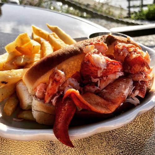 A lobster roll filled with chunks of lobster meat, served with a side of golden fries on a sunny outdoor table.