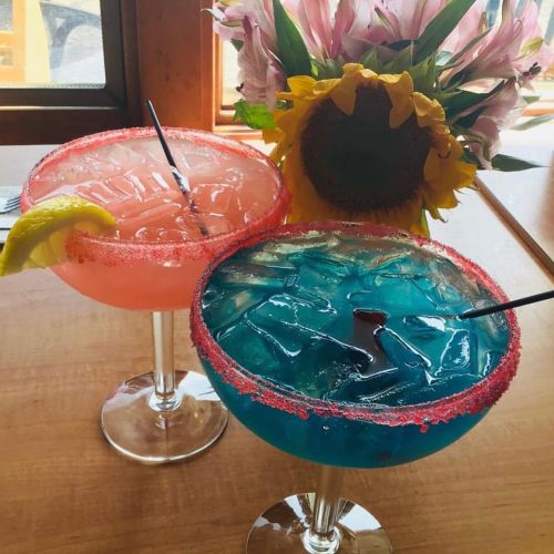 Two colorful cocktails in large glasses, one pink with a lemon slice and one blue, beside a sunflower and pink flowers.