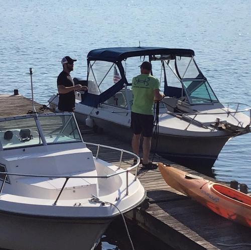 Two men on a dock near boats, one preparing equipment while the other stands by a kayak on the water.