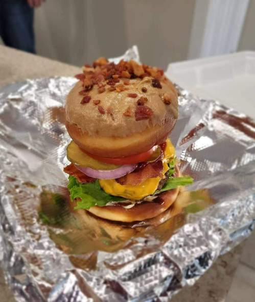 A towering burger with a donut bun, layered with lettuce, tomato, cheese, and bacon, wrapped in foil.