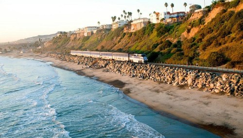 A train travels along a coastal track beside a sandy beach and rocky shoreline, with palm trees and cliffs in the background.