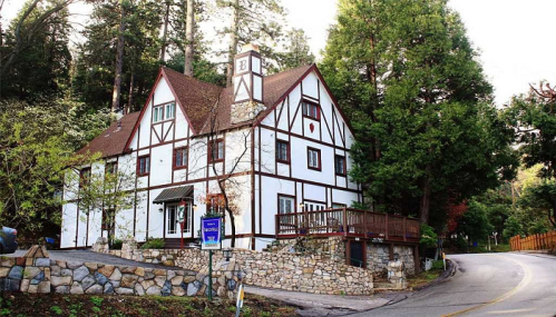A charming, two-story Tudor-style house with a stone wall, surrounded by trees and a winding road.