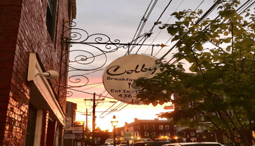 Sign for Colby's Breakfast with a sunset backdrop, surrounded by trees and power lines.