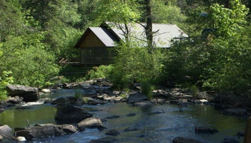 A serene cabin by a stream, surrounded by lush greenery and trees in a peaceful natural setting.