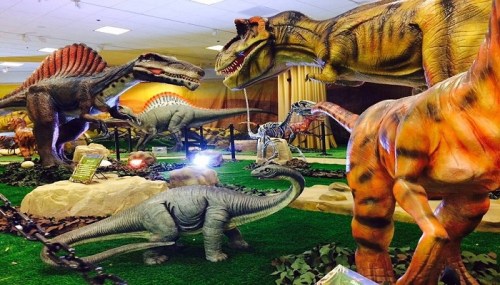 A vibrant display of life-sized dinosaur models in a museum setting, featuring various species and lush greenery.