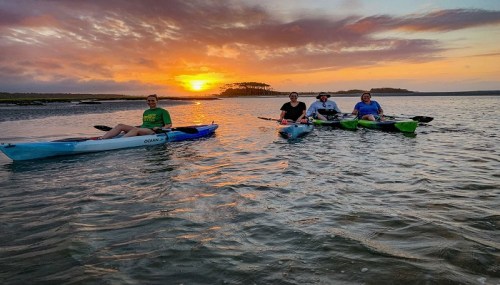 Four people kayaking on a calm waterway at sunset, with vibrant colors in the sky and silhouettes of trees in the background.