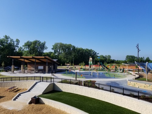A sunny park featuring a splash pad, playground equipment, and picnic areas surrounded by greenery.