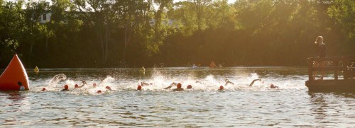 A group of swimmers in orange caps splashes through a sunlit lake, with a person observing from a dock.