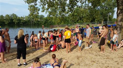 A busy beach scene with children and adults enjoying the water, playing, and relaxing on the sand under trees.
