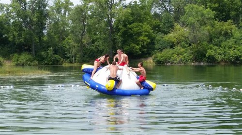 Three young men play on a large inflatable water toy in a lake, surrounded by trees and calm water.
