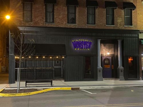 Exterior of a restaurant named "Famous Monster Pizza," featuring a dark facade and illuminated sign at night.