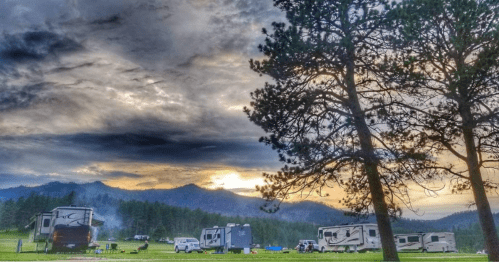 A scenic campsite with RVs under a dramatic sky, surrounded by trees and mountains at sunset.