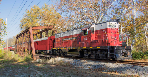 A vintage red train crosses an old metal bridge surrounded by autumn trees.