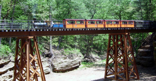 A colorful train crosses a wooden bridge over a rocky gorge, surrounded by lush green trees.