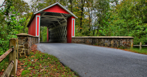 A red covered bridge with a stone entrance, surrounded by lush greenery and a paved road leading through it.