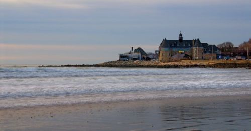 A calm beach scene with gentle waves and a historic stone building in the background under a cloudy sky.