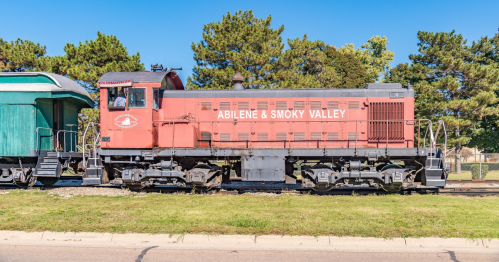 A red and black diesel locomotive labeled "Abilene & Smoky Valley" parked beside a green train car on a sunny day.