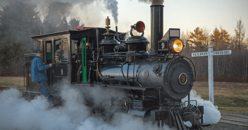 A vintage steam locomotive with a conductor, billowing smoke, near a railroad crossing sign in a scenic landscape.