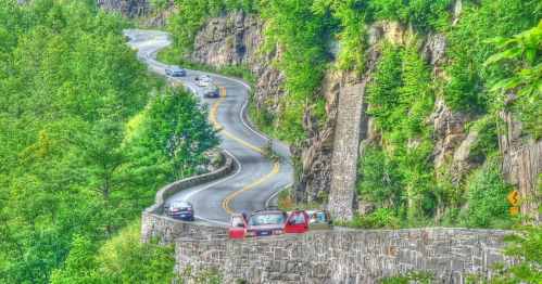 Winding road through lush greenery with cars parked along a stone wall, surrounded by rocky cliffs.