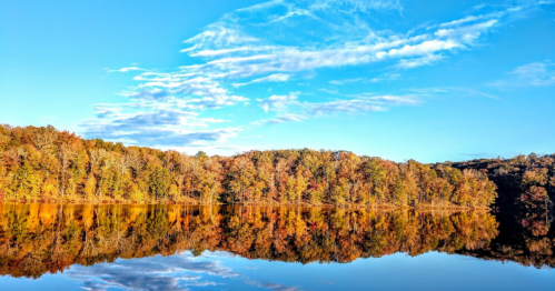 A serene lake reflecting vibrant autumn trees under a clear blue sky with scattered clouds.