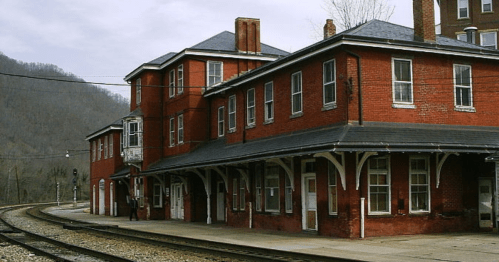 Historic red brick train station with a curved platform, set against a backdrop of rolling hills and cloudy skies.