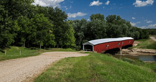 A red covered bridge beside a river, surrounded by lush green trees and a clear blue sky.