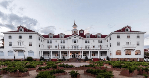 Historic white hotel with a red roof and tower, surrounded by greenery and a cloudy sky.