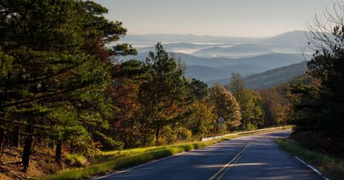 A winding road through a scenic landscape with mountains and trees, under a clear sky.