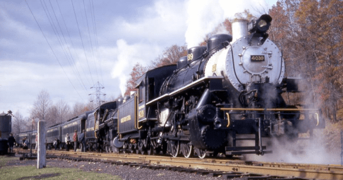 Two vintage steam locomotives at a station, with smoke billowing and autumn foliage in the background.